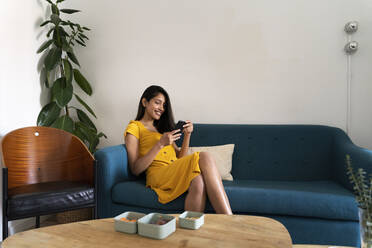 Smiling young woman sitting on couch using cell phone - AFVF04477
