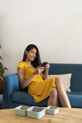 Lächelnde junge Frau sitzt auf der Couch und benutzt ein Mobiltelefon - AFVF04478