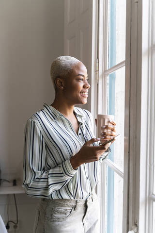 Glückliche Frau mit Handy und Kaffeetasse schaut aus dem Fenster, lizenzfreies Stockfoto