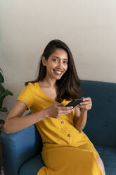 Porträt einer lächelnden jungen Frau, die auf einer Couch sitzt und ein Mobiltelefon hält - AFVF04516