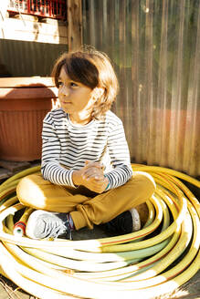 Porträt eines entspannten Jungen, der zwischen aufgerollten Gartenschläuchen in einer Gärtnerei sitzt - VABF02489