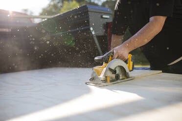 Midsection of man using electric saw while sawing wooden plank in yard - CAVF70787