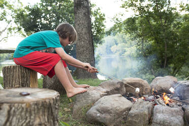 Seitenansicht eines Jungen, der auf einem Baumstumpf auf dem Campingplatz sitzend Marshmallows über einem Lagerfeuer röstet - CAVF70865