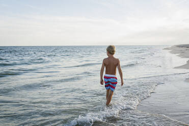Rückansicht eines Jungen ohne Hemd, der am Tobay Beach in den Wellen gegen den Himmel läuft - CAVF70876