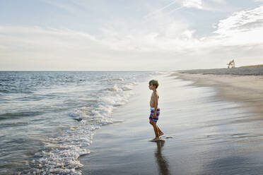 Side view of shirtless boy looking away while standing at Tobay Beach against sky - CAVF70878