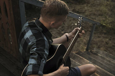 Hoher Blickwinkel eines Mannes, der Gitarre spielt, während er auf den Stufen einer Veranda sitzt - CAVF70899