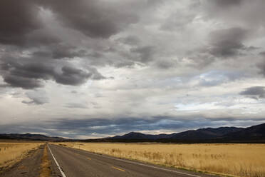 Landstraße inmitten von Feldern gegen stürmische Wolken und Berge - CAVF70949