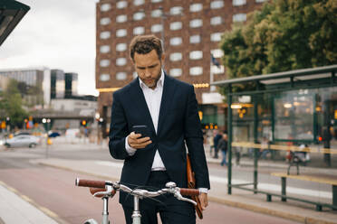 Geschäftsmann, der ein Mobiltelefon benutzt, während er mit dem Fahrrad auf einer Straße in der Stadt steht - MASF15515