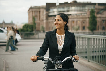 Lächelnde Geschäftsfrau mit Fahrrad auf einer Brücke in der Stadt - MASF15516