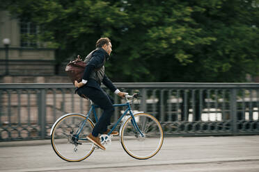 Seitenansicht eines Geschäftsmannes mit Tasche, der auf einer Brücke Fahrrad fährt - MASF15523