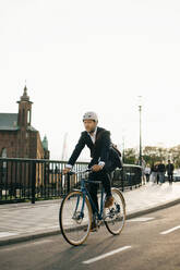 Confident businessman riding bicycle on street in city against clear sky - MASF15535