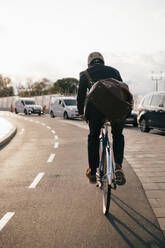 Rear view of businessman riding bicycle on street in city during sunny day - MASF15537