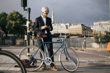 Geschäftsmann, der ein Mobiltelefon benutzt, während er mit dem Fahrrad auf einer Straße in der Stadt steht - MASF15539