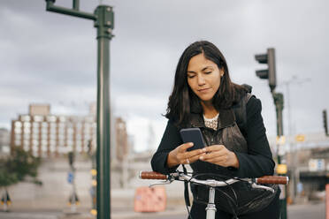 Geschäftsfrau, die ein Mobiltelefon benutzt, während sie sich in der Stadt auf einen Fahrradgriff stützt - MASF15545