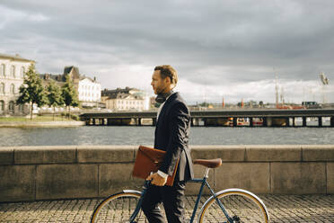 Seitenansicht von Geschäftsmann zu Fuß mit Fahrrad auf Gehweg in der Stadt gegen Himmel - MASF15556