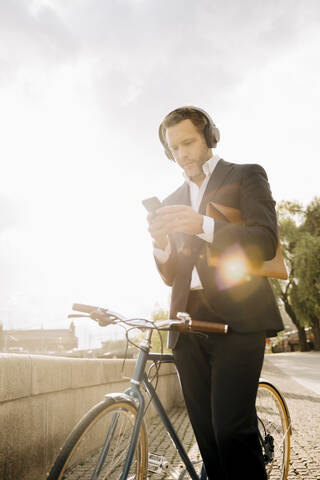 Geschäftsmann, der ein Mobiltelefon benutzt, während er mit dem Fahrrad gegen den Himmel an einem sonnigen Tag steht, lizenzfreies Stockfoto