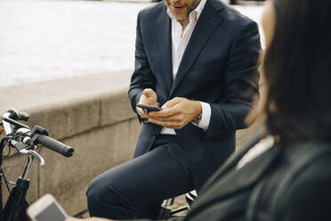 Mittelteil eines Geschäftsmannes, der ein Mobiltelefon benutzt, während er auf einem Fahrrad sitzt, mit einer Geschäftsfrau - MASF15565