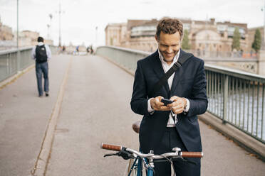 Smiling businessman using mobile phone while standing with bicycle on bridge in city - MASF15576