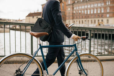 Mittelteil eines Geschäftsmannes, der ein Smartphone benutzt, während er mit dem Fahrrad auf einer Brücke in der Stadt spazieren geht - MASF15578