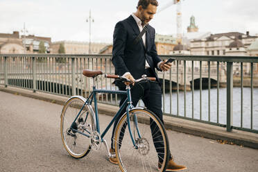 Confident businessman using smart phone while walking with bicycle on bridge in city - MASF15579