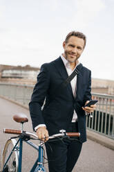 Portrait of confident businessman holding mobile phone while walking with bicycle on bridge in city - MASF15582