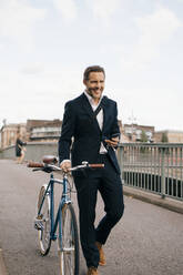 Smiling businessman holding smart phone while walking with bicycle on bridge in city - MASF15583