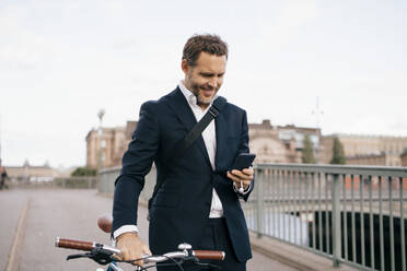 Smiling businessman using mobile phone while walking with bicycle on bridge in city - MASF15584