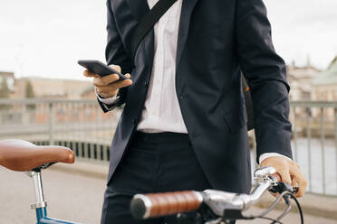 Midsection of businessman using smart phone while standing with bicycle on bridge in city - MASF15586
