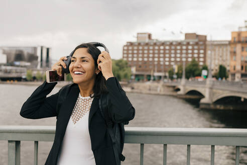 Lächelnde Geschäftsfrau mit Kopfhörern, die auf einer Brücke in der Stadt steht - MASF15587