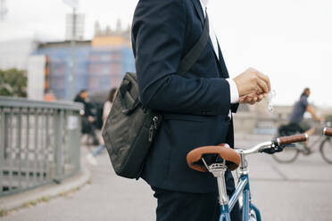 Midsection of businessman holding in-ear headphones while standing with bicycle on bridge in city - MASF15589