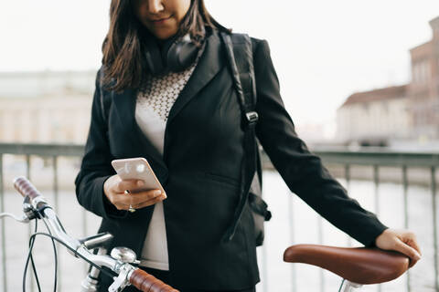 Mittelteil einer Geschäftsfrau, die ein Mobiltelefon benutzt, während sie mit dem Fahrrad auf einer Brücke in der Stadt steht, lizenzfreies Stockfoto