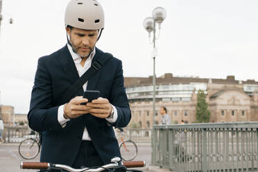 Geschäftsmann, der ein Mobiltelefon benutzt, während er mit dem Fahrrad auf einer Brücke in der Stadt steht - MASF15599