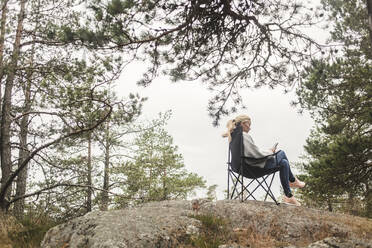 Full length of woman using mobile phone while sitting on camping chair over rock formation against trees - MASF15602