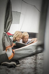 Lächelnde Geschwister, die ein digitales Tablet benutzen, während sie im Zelt auf dem Campingplatz liegen - MASF15617