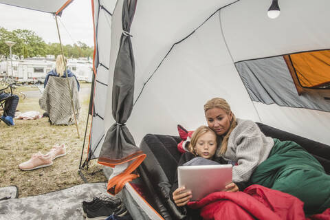Mädchen schaut auf ein digitales Tablet, während sie mit ihrer Mutter im Zelt auf dem Campingplatz liegt, lizenzfreies Stockfoto
