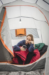 Teenage girl using smart phone while sitting in tent at camping site - MASF15624