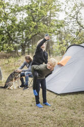Junge nimmt Schwester mit Smartphone huckepack, während Mann mit Hund beim Zelten spielt - MASF15634