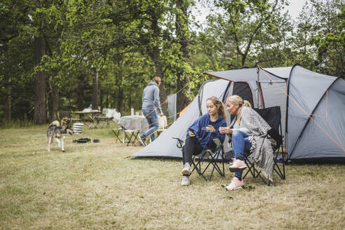 Mutter und Tochter trinken Kaffee, während sie auf dem Campingplatz auf ihr Mobiltelefon schauen - MASF15637