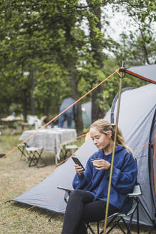 Teenager-Mädchen, das ein Mobiltelefon benutzt, während es Kaffee gegen ein Zelt trinkt - MASF15639