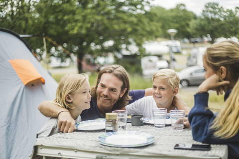 Lächelnder Vater sitzt mit Kindern am Tisch auf dem Campingplatz, lizenzfreies Stockfoto