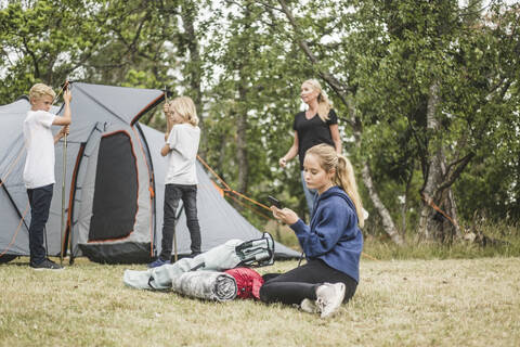 Teenager text messaging on mobile phone while family pitching tent stock photo