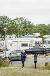 Kinder, die gemeinsam eine aufblasbare Matratze tragen, während sie auf dem Campingplatz gegen einen Wohnwagen laufen - MASF15676