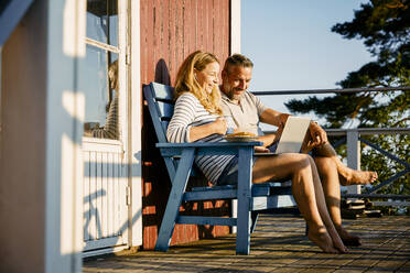 Smiling couple using laptop while having breakfast at porch in log cabin - MASF15692