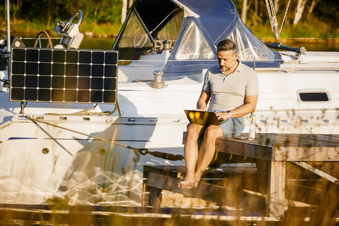 Mature man working on laptop while sitting at pier against yacht over lake - MASF15699