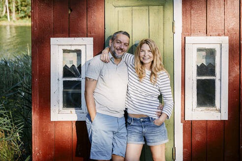 Porträt eines lächelnden Paares, das mit umschlungenen Armen vor der Tür einer Blockhütte steht - MASF15705