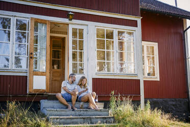 Paar schaut auf sein Smartphone, während es auf den Stufen vor der Blockhütte sitzt - MASF15707