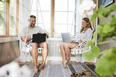 Lächelndes Paar benutzt Laptops, während es auf einer Seilschaukel in einer Blockhütte sitzt - MASF15715