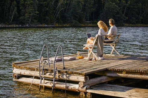 Man and woman using laptop while sitting on bench over pier against lake during summer - MASF15718