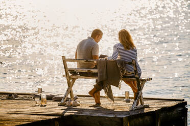 Full length of couple using laptop while sitting on bench over wooden pier against lake during summer vacation - MASF15720