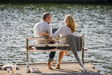Rückansicht eines Paares, das sich auf einer Bank am Seeufer mit einem Laptop unterhält - MASF15721